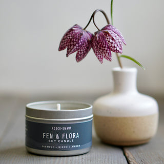 A Rosco-Emmit FEN & FLORA soy candle in a silver tin with a slate blue label, styled on a rustic wooden surface next to a ceramic vase holding purple-and-white checkered flowers.