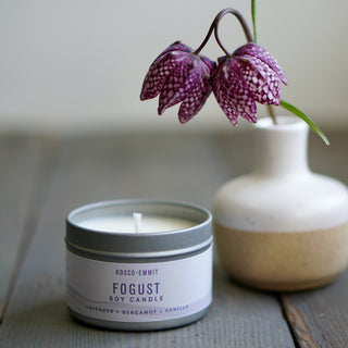 A candle tin labeled ROSCO-EMMIT FOGUST SOY CANDLE sits on a rustic wooden surface, with a ceramic vase holding a purple-and-white spotted flower behind it.