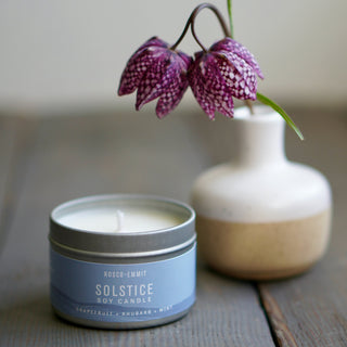 A Solstice soy candle in a silver tin with a light blue label sits on a rustic wooden surface next to a small ceramic vase holding purple-and-white checkered flowers.