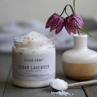 A glass jar of Rosco-Emmit Cedar Lavender Bubble Salts sits on a rustic wooden surface, with a small wooden spoon holding some of the white salt crystals in front of it. A ceramic vase with a purple-and-white hanging flower is blurred in the background.