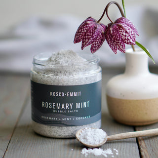 A jar of Rosco-Emmit Rosemary Mint Bubble Salts sits on a rustic wooden table, with a small wooden spoon holding white salt in front and a ceramic vase with hanging purple flowers behind it.