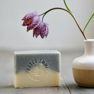 A rectangular bar of layered soap standing on a wooden surface, with a white ceramic vase and a hanging purple flower in the background. The soap is embossed with the brand name Rosco Emmet and Vancouver Island.