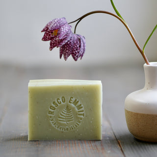 A rectangular bar of pale yellow-green soap with a circular embossed logo reading 'Rosco Emma' and 'Malcooem Island' sits on a rustic wooden surface. A small white ceramic vase with a purple spotted flower is beside it, and the background is softly blurred.