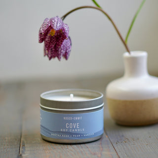 A Rosch-Emmitt 'Cove' soy candle in a silver tin with a light blue label, styled on a rustic wooden table with a purple flower and a ceramic vase in the background.