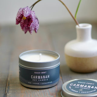 An open silver tin candle labeled ROSCO-EMMIT CARMANAH soy candle sits on a rustic wooden surface, with a matching lid placed beside it and a small white-and-beige vase holding a purple spotted flower in the background.