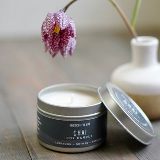 An open silver tin candle labeled ROSCO-EMMIT CHAI SOY CANDLE sits on a wooden surface with its lid leaning beside it. A white ceramic vase with a hanging pink-purple flower is blurred in the background.