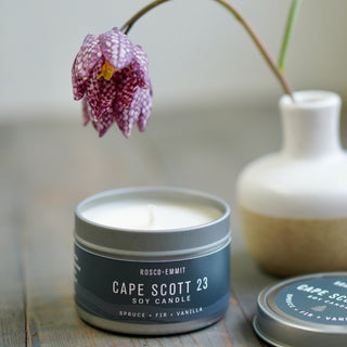 A tin candle labeled ROSCO-EMMIT CAPE SCOTT 23 SOY CANDLE sits on a wooden surface, with a white ceramic vase and a hanging purple flower blurred in the background.