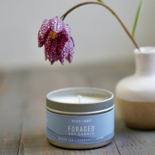 A candle tin labeled ROSCO-EMMIT FORAGED SOY CANDLE sits on a rustic wooden surface, with a purple spotted flower hanging above it and a small white vase blurred in the background.