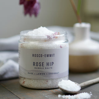 A clear glass jar of Rosco-Emmit Rose Hip Bubble Salts filled with white salt crystals, set on a rustic wooden surface with a wooden spoon of salt in the foreground and a blurred ceramic vase in the background.