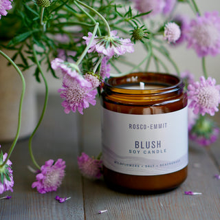 An amber glass soy candle jar labeled ROSCO-EMMIT BLUSH SOY CANDLE, styled with pink wildflowers on a rustic wooden surface.