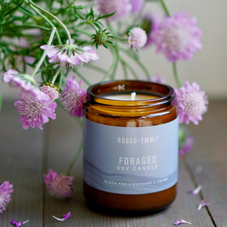 An amber glass jar candle labeled ROSCO-EMMIT Foraged Soy Candle sits on a wooden surface surrounded by soft pink flowers. The candle is shown in a styled natural setting with no people or animals.