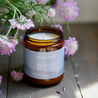 An amber glass soy candle jar with a pale blue label reading ROSCO-EMMIT SOLSTICE SOY CANDLE, styled on a rustic wooden surface with pink flowers around it.