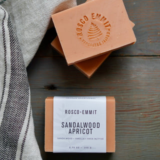 Two bars of Rosco-Emmit Sandalwood Apricot handmade soap are displayed on a rustic dark wood background with a gray striped cloth. One bar is stacked on top of the other, showing the embossed brand logo, while the other is wrapped in a white paper band with product details.