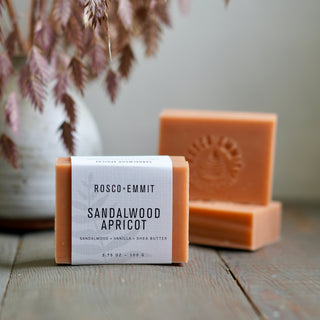 A bar of Rosco-Emmit Sandalwood Apricot soap in its paper wrapper, displayed on a rustic wooden surface with a second unwrapped bar of soap behind it and dried botanicals in the background.