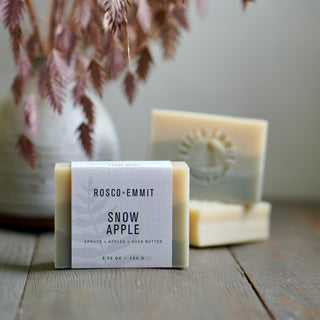 A bar of soap labeled 'ROSCO-EMMIT SNOW APPLE' sits on a rustic wooden surface, with two additional soap bars blurred in the background near a vase of dried pink branches.
