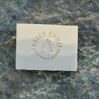 A rectangular translucent white soap bar embossed with 'ROSCO EMMIT VANCOUVER ISLAND' and a pine tree logo, placed on a textured gray stone surface.