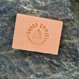 A rectangular bar of peach-colored soap with an embossed circular logo reading 'Rosco Emmett Vancouver Island' and a fern design, placed on a textured gray rock surface.