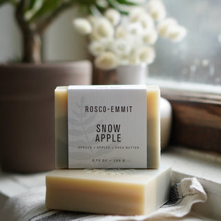 A bar of Rosco-Emmit Snow Apple soap stacked on another bar, with a white paper label wrapped around the front. The soap is photographed on a soft fabric surface near a window, with blurred potted plants and white flowers in the background.
