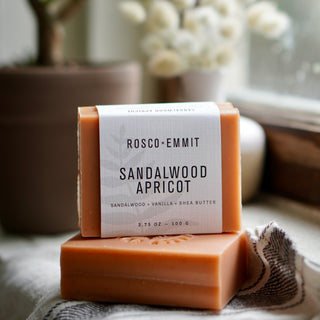 A bar of Rosco Emmit Sandalwood Apricot handmade conditioner soap stacked on another bar, with a white paper label, set on a textured cloth with a blurred background.