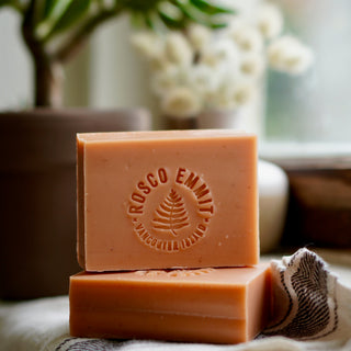Two rectangular bars of Rosco Enlit Margocolloid soap stacked on a cloth, with a potted plant and white flowers blurred in the background.