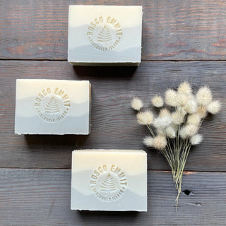 Three rectangular bars of Rosco Emily handcrafted soap with a mountain landscape embossed on the top, arranged on a dark rustic wooden background next to a small bunch of dried bunny tail grass.