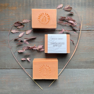 Three bars of Rosco Emiss Sandalwood Apricot soap arranged on a rustic gray wooden surface, with two bars embossed and one wrapped in a white paper label. Dried pinkish-brown grasses are arranged around the bars in a decorative flat-lay composition.