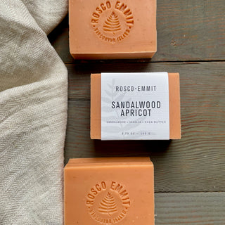 Three bars of Rosco-Emmit Sandalwood Apricot soap arranged on a rustic wooden surface with a beige linen cloth. The center bar has a white paper label, while the top and bottom bars are embossed with the brand name.